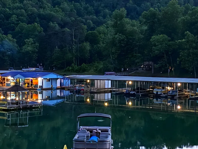 Boat dock with lights reflecting on calm lake at dusk, surrounded by trees.